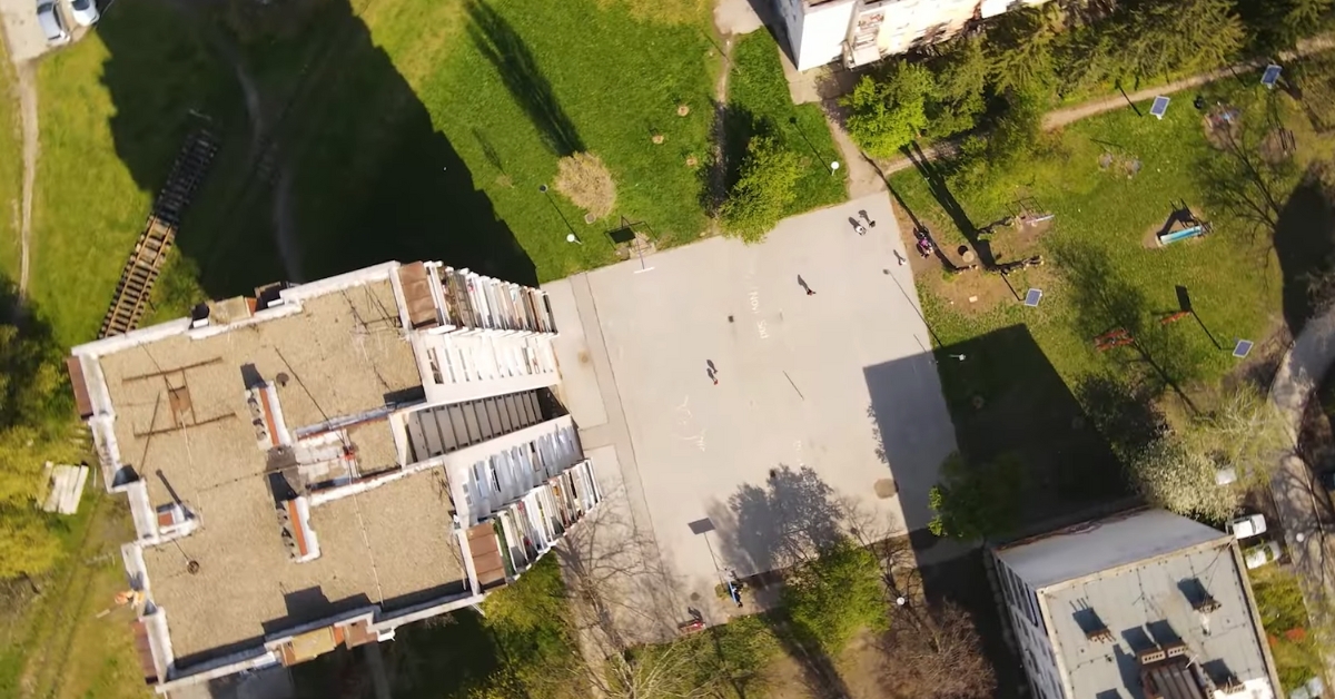 Pogled iz vazduha na mirno dvorište između zgrada u Novom Sadu, sa zelenilom i nekoliko dece na betonskom igralištu